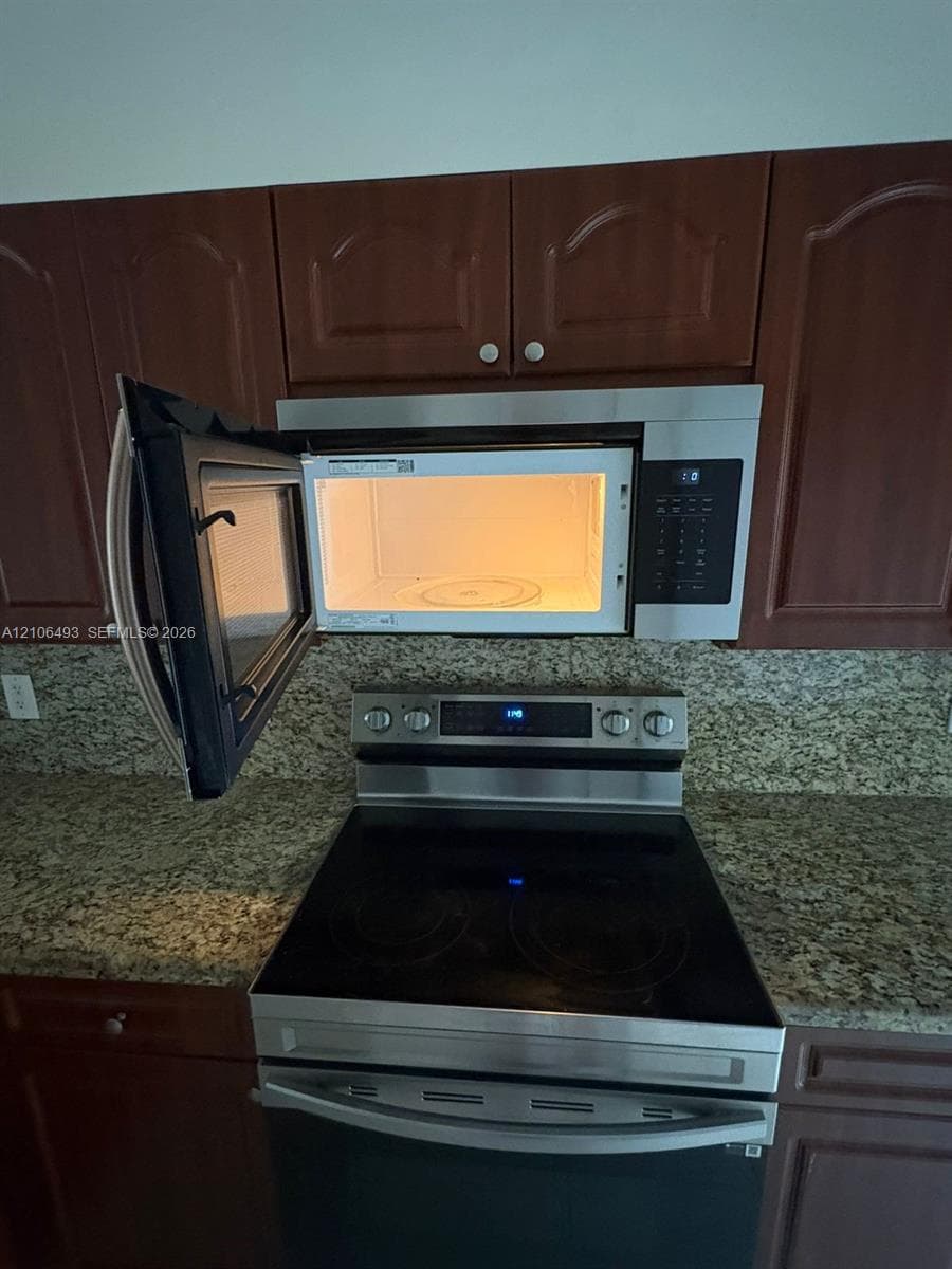 Stainless steel over-the-range microwave and electric range, dark wood-finish cabinetry, and stone-surface countertops with matching backsplash