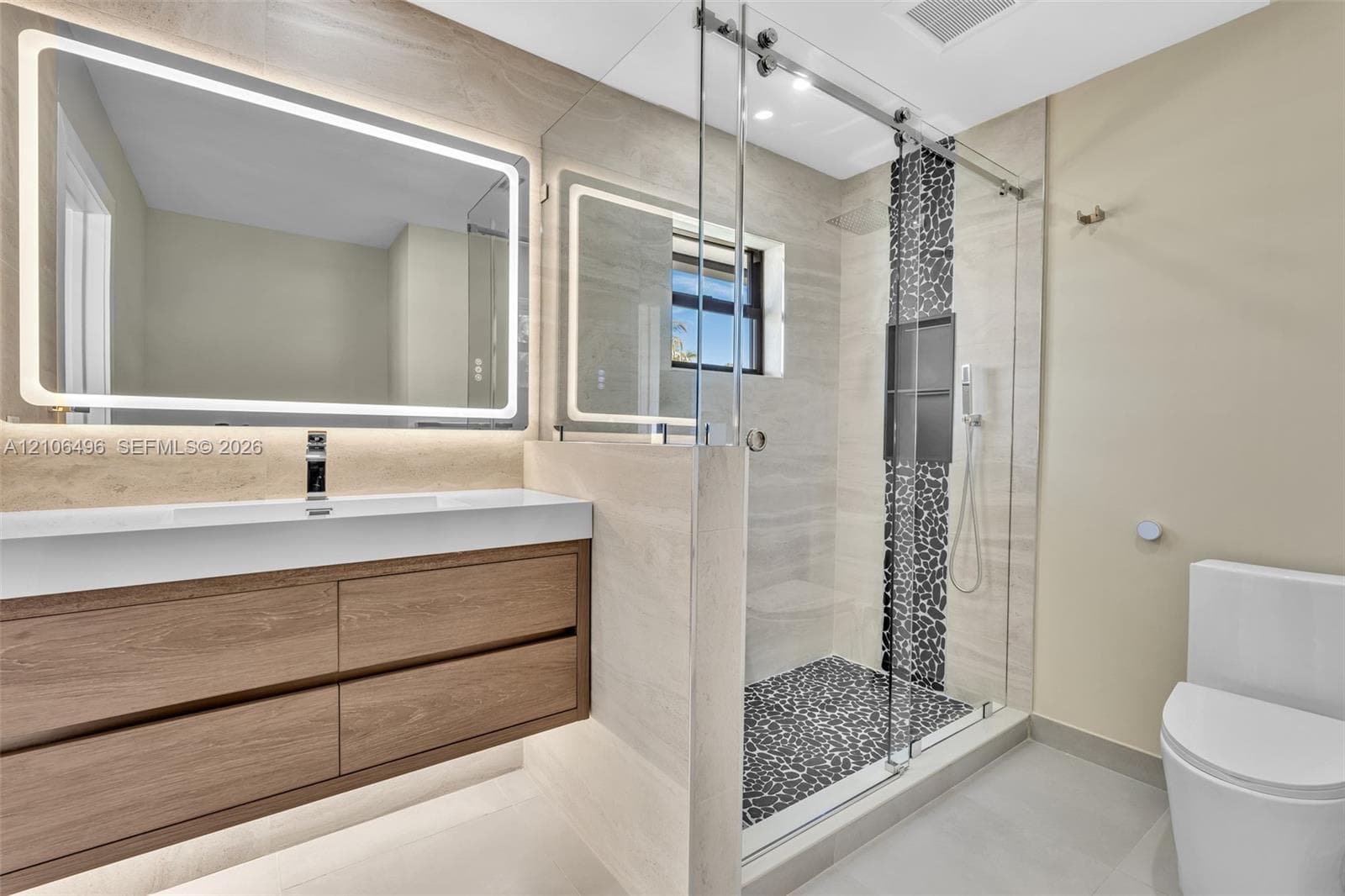 Floating wood-finish vanity with an integrated countertop, large LED-backlit mirror, and walk-in shower featuring glass enclosure, pebble stone flooring, and recessed shelving