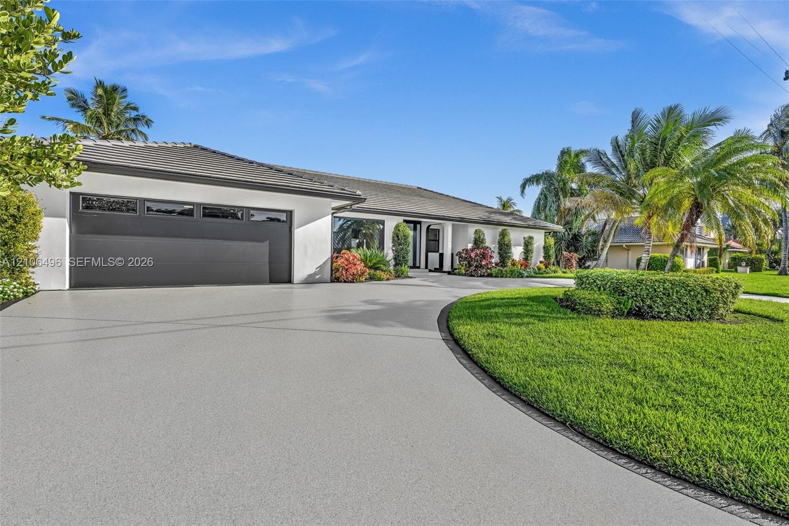 Single-story exterior featuring white stucco siding, a multi-car garage with overhead glass-paneled door, a curved concrete driveway, and mature landscaping