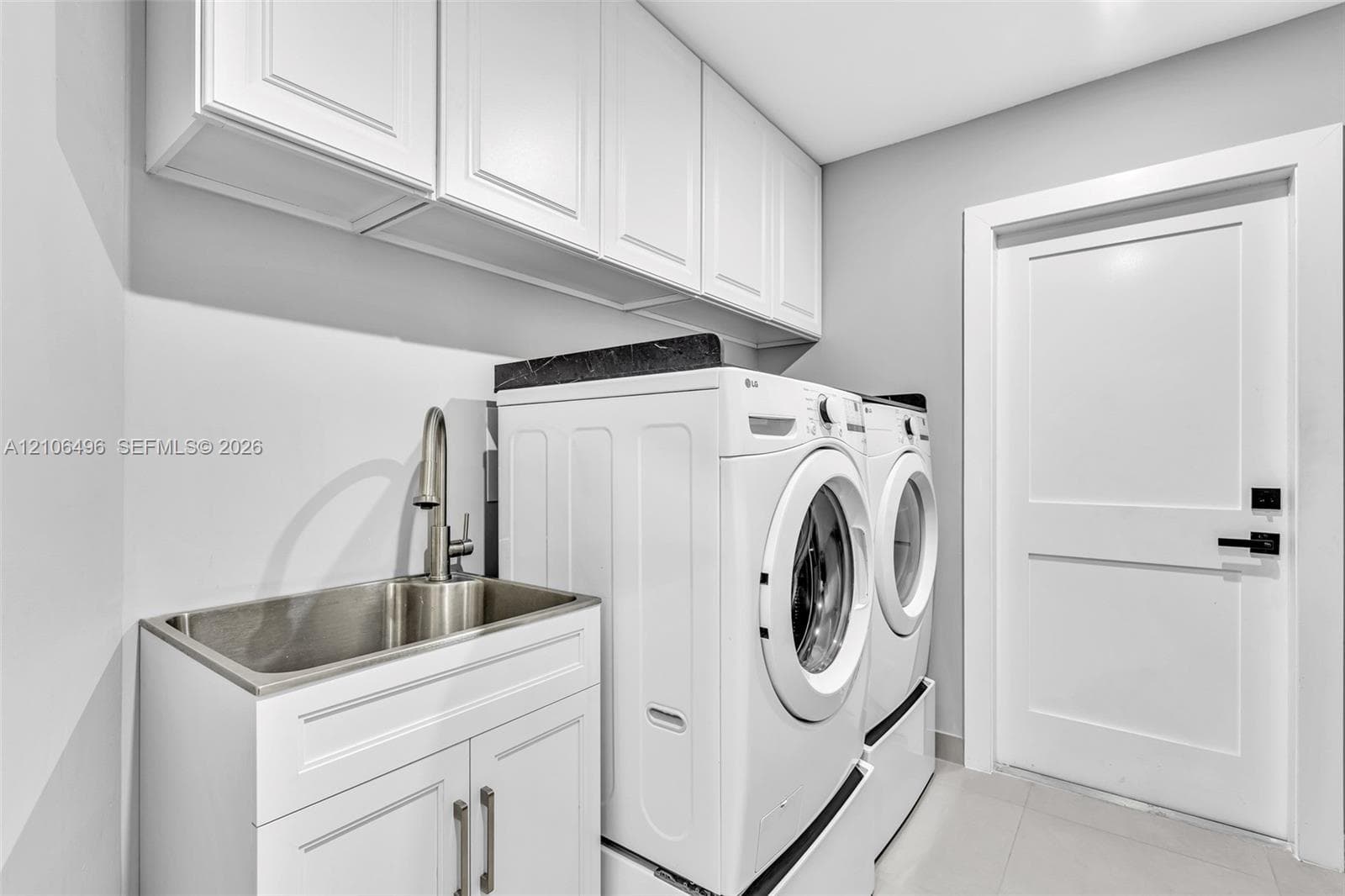 Laundry room featuring white shaker-style upper and lower cabinetry, an integrated stainless steel utility sink with a high-arch pull-down faucet, dark stone-finish countertop, and a solid-core door with matte black hardware