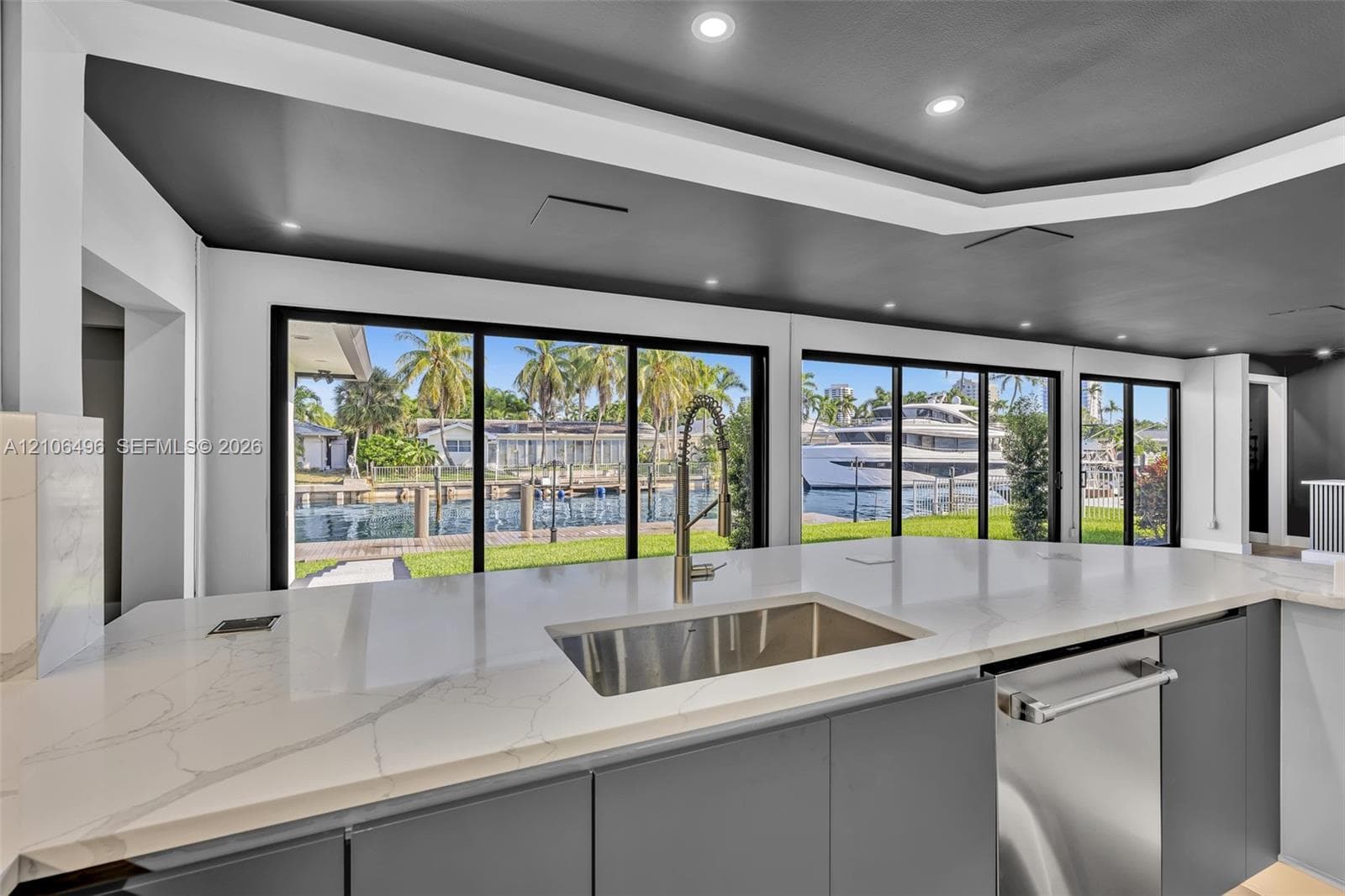 Large kitchen island with white countertop, undermount stainless steel sink, gooseneck faucet, gray flat-panel cabinetry, and extensive sliding glass doors