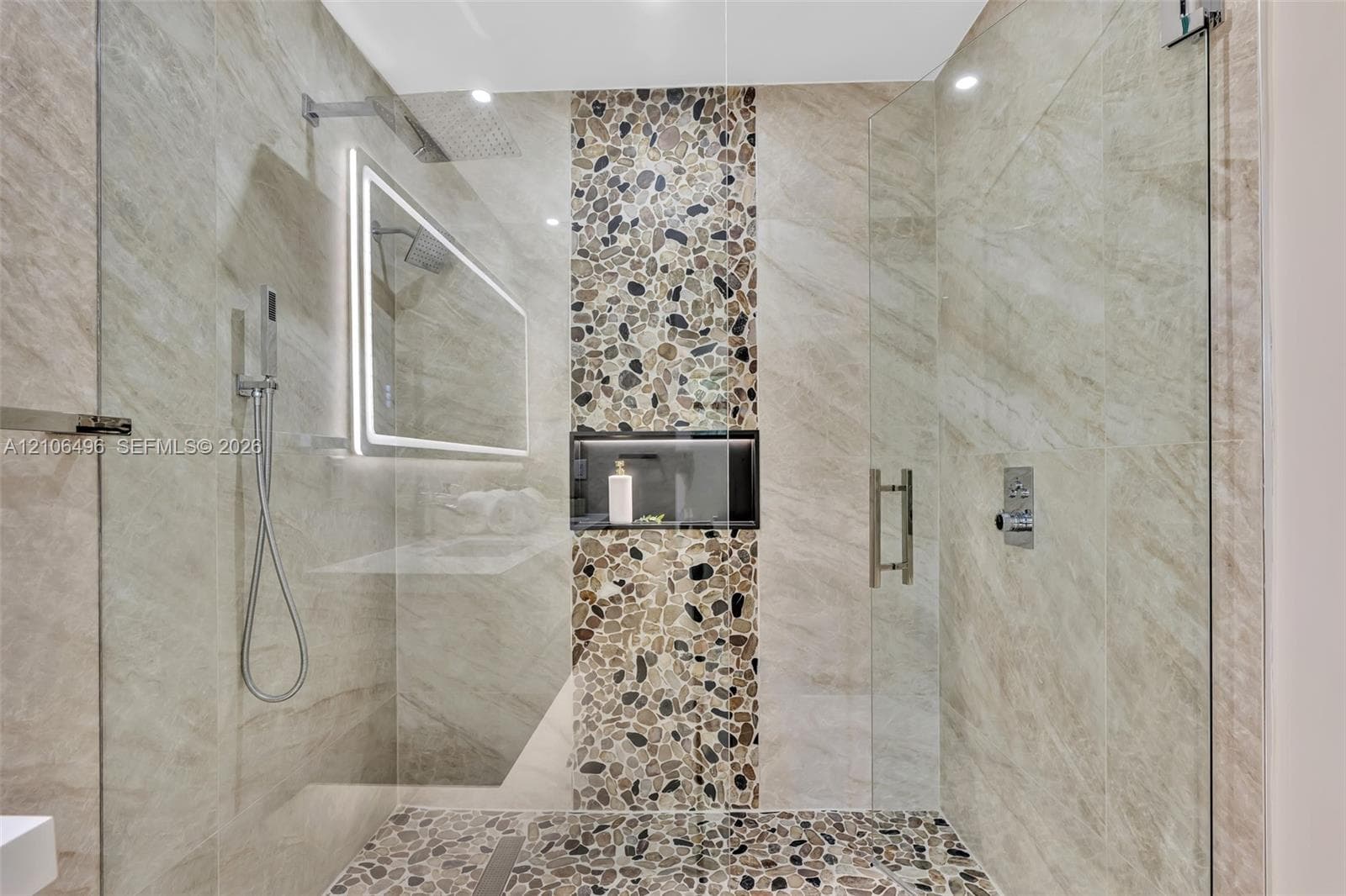 Walk-in shower featuring large-format neutral wall tile, a vertical pebble-stone accent column, pebble-tile flooring, a recessed storage niche, and an LED-backlit mirror