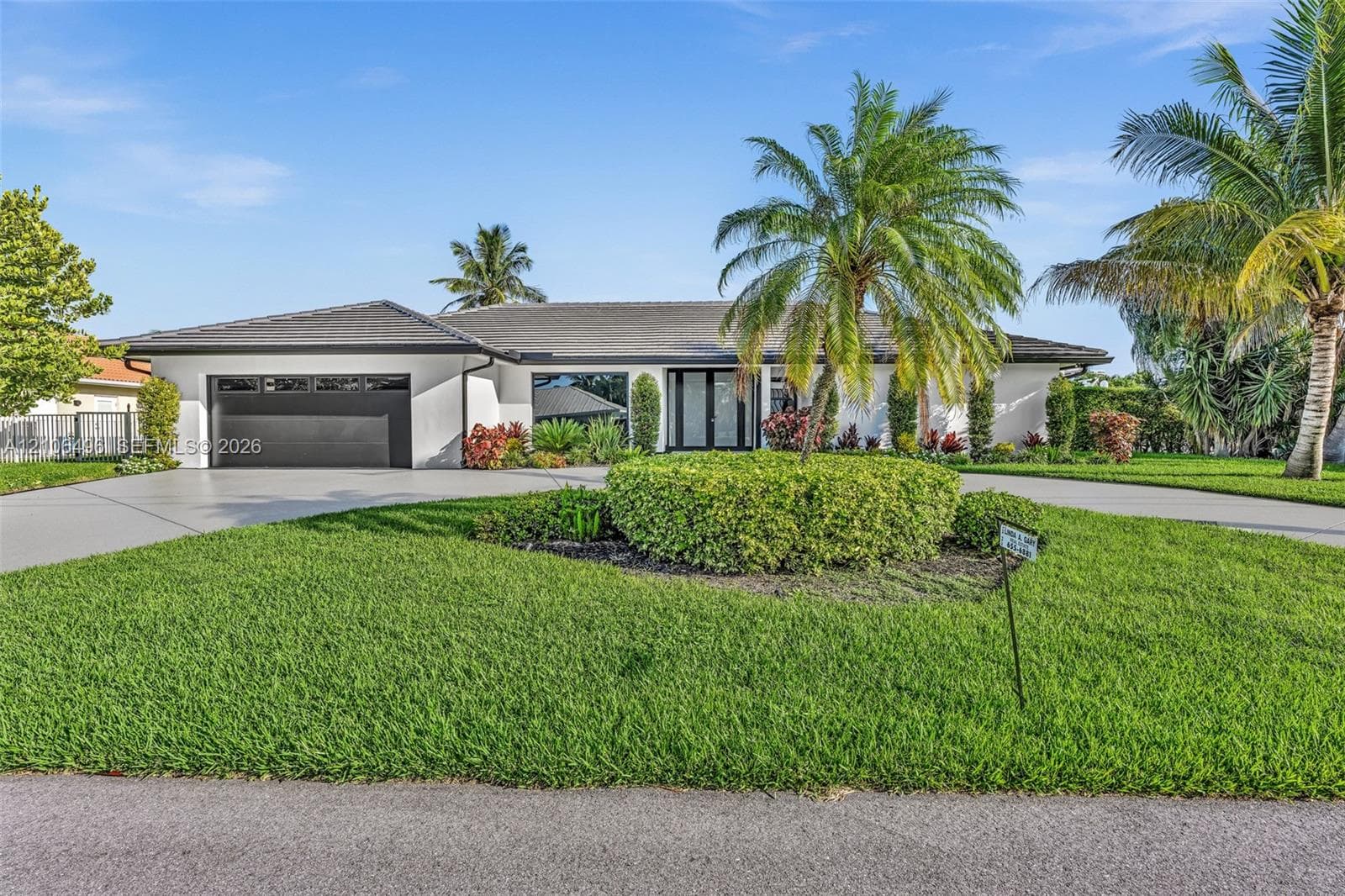 Single-story residence featuring a smooth stucco facade, hip roof with dark tiles, attached two-car garage, and manicured landscaping with established palm trees
