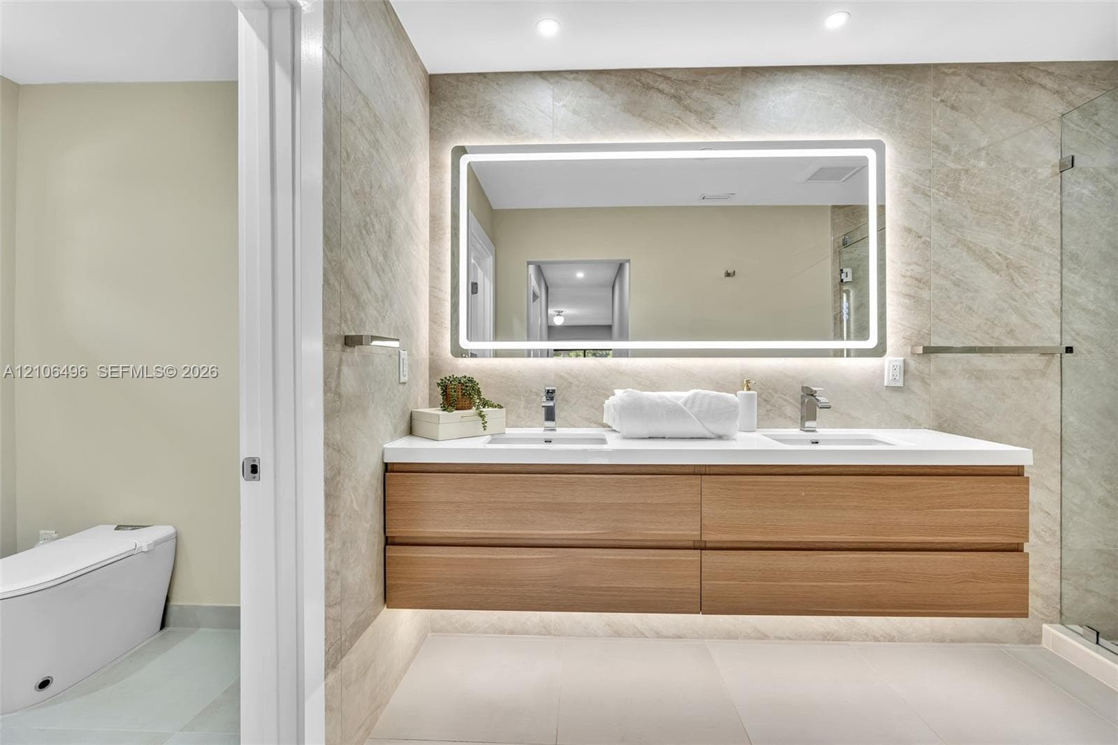 Floating wood-finish vanity with dual integrated sinks, quartz countertop, illuminated LED vanity mirror, full-height stone-finish wall tile, and glass-enclosed shower