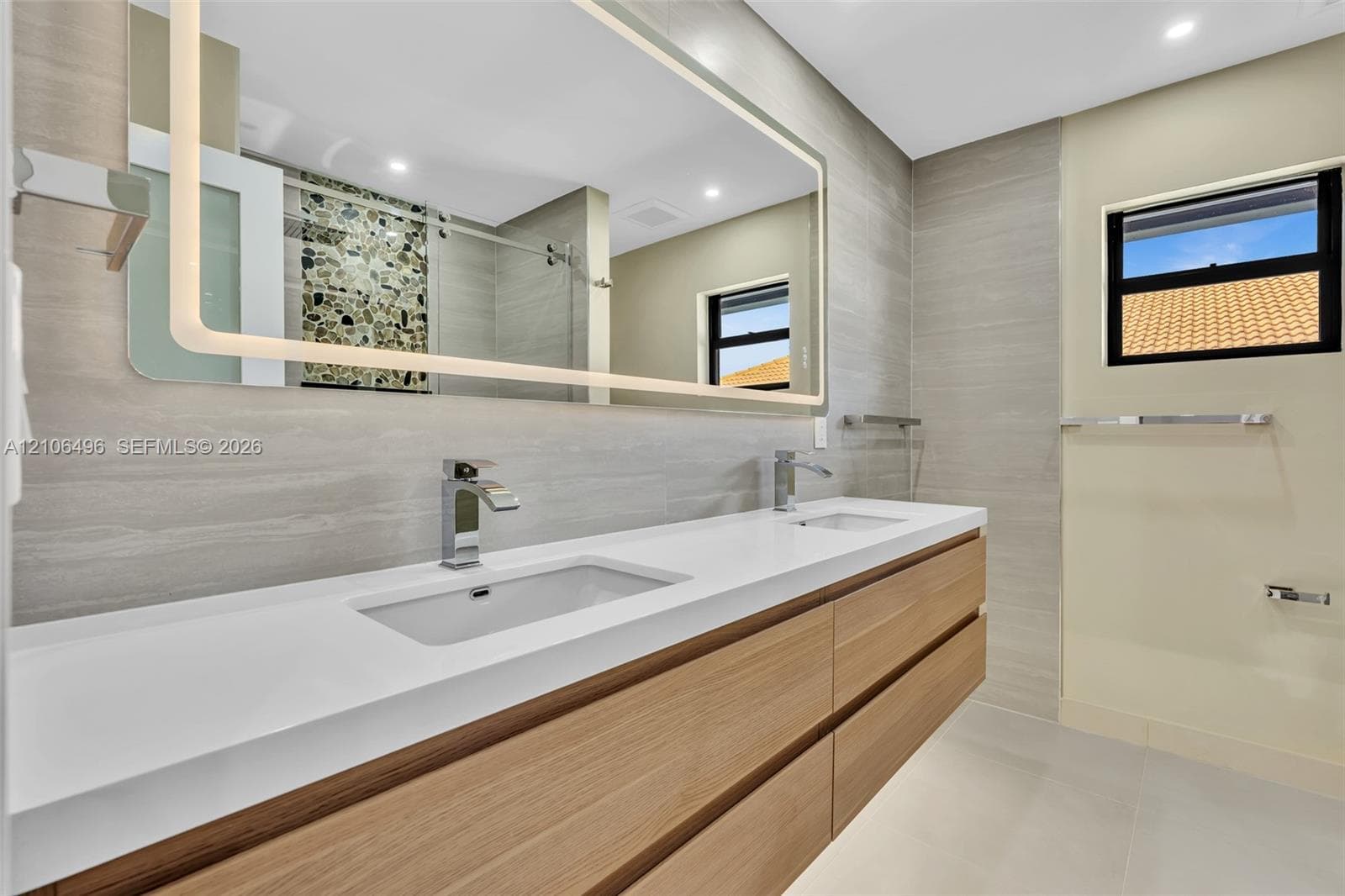 Double vanity featuring a white composite countertop, integrated rectangular sinks, wood-finish cabinetry, a large back-lit mirror, and neutral wall-tile surround