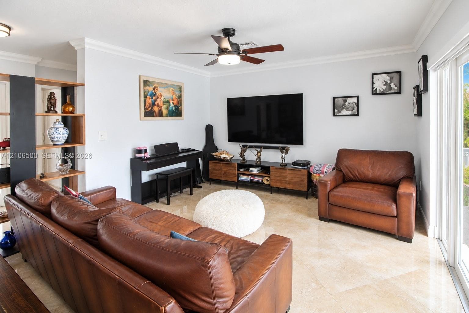 Living area featuring large-format light-toned flooring, crown molding, built-in wooden shelving, and a ceiling fan with integrated lighting