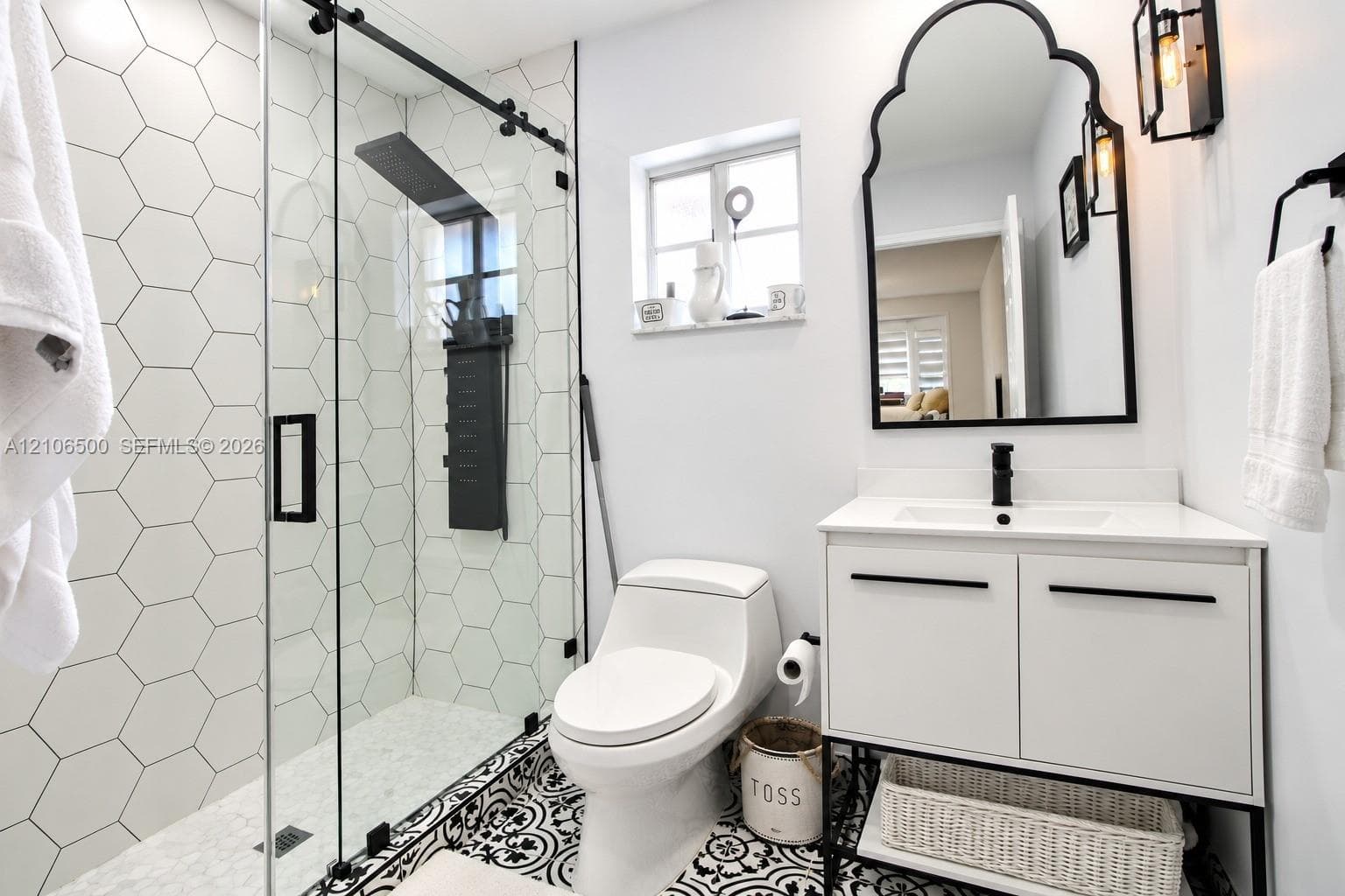 Walk-in shower with hexagon wall tile, sliding glass door, and rainfall showerhead; vanity featuring a rectangular sink, matte black hardware, and an arched mirror
