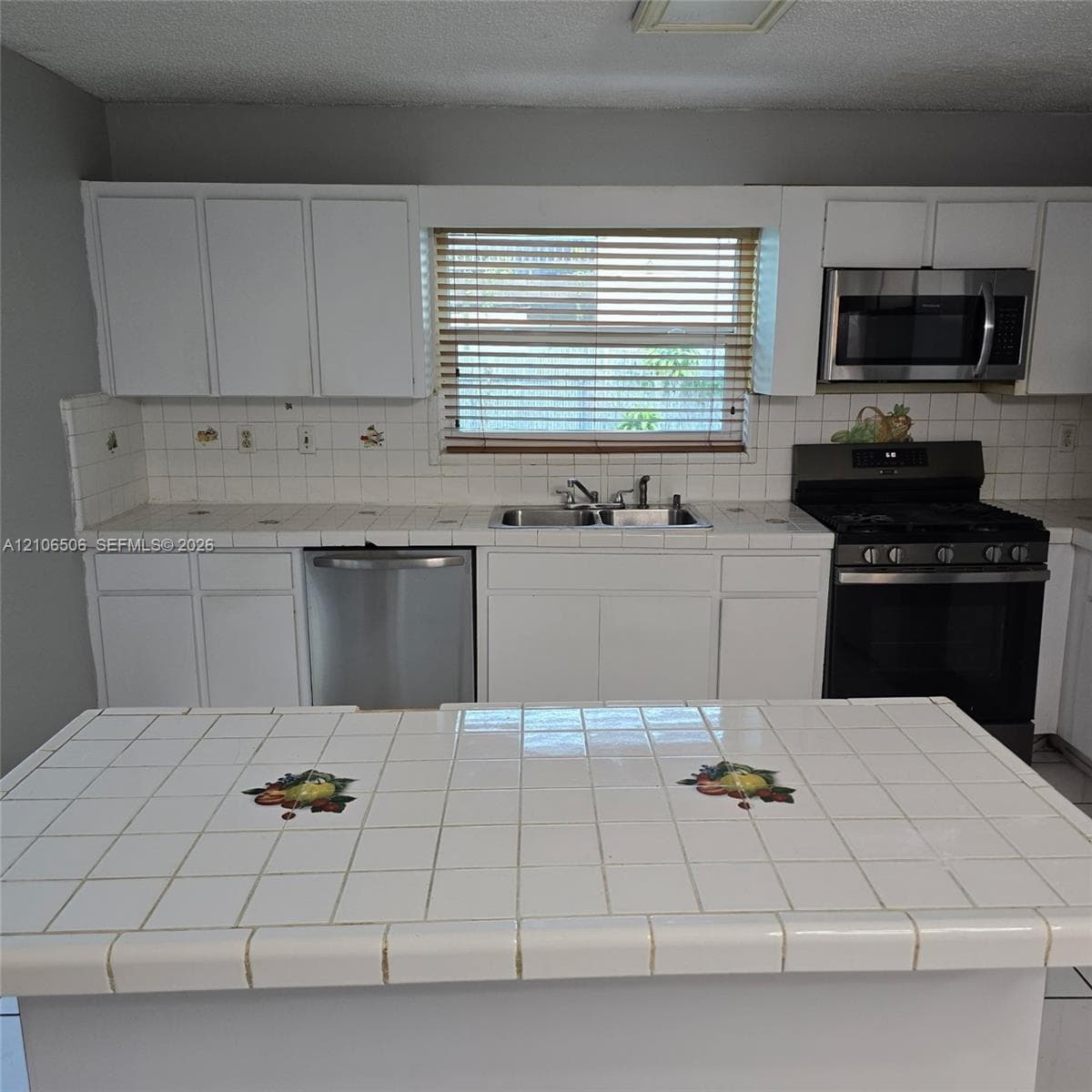 Kitchen featuring white cabinetry, tiled countertops with decorative inlays, a stainless steel dishwasher, an integrated over-the-range microwave, and a stainless steel gas range