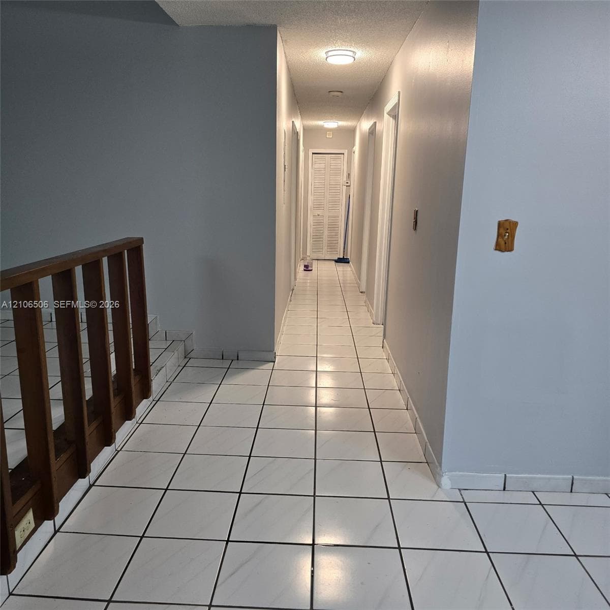 Hallway featuring ceramic tile flooring, a wood-finish staircase railing, and louvered closet doors