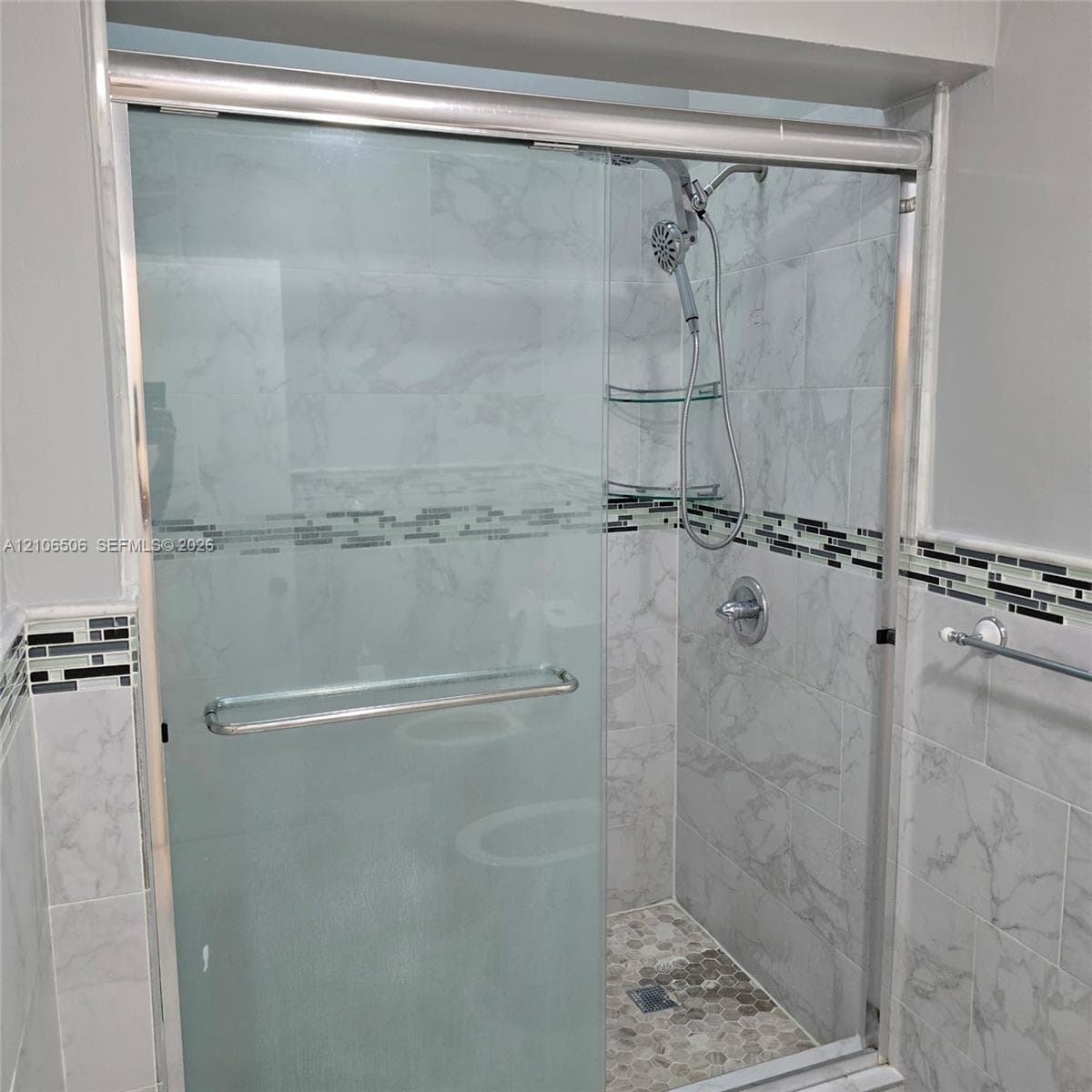Walk-in shower enclosure featuring large-format stone-patterned wall tile, horizontal glass-mosaic accent trim, a pebble-finish floor, and a handheld shower fixture