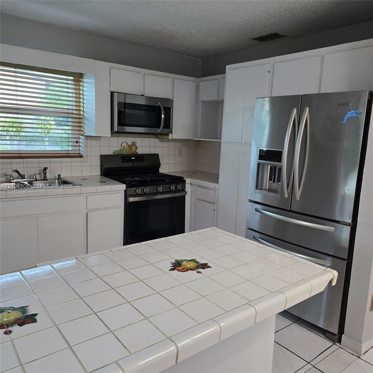 Kitchen featuring white cabinetry, tiled countertops with decorative inlays, stainless steel refrigerator, integrated microwave, and a gas range