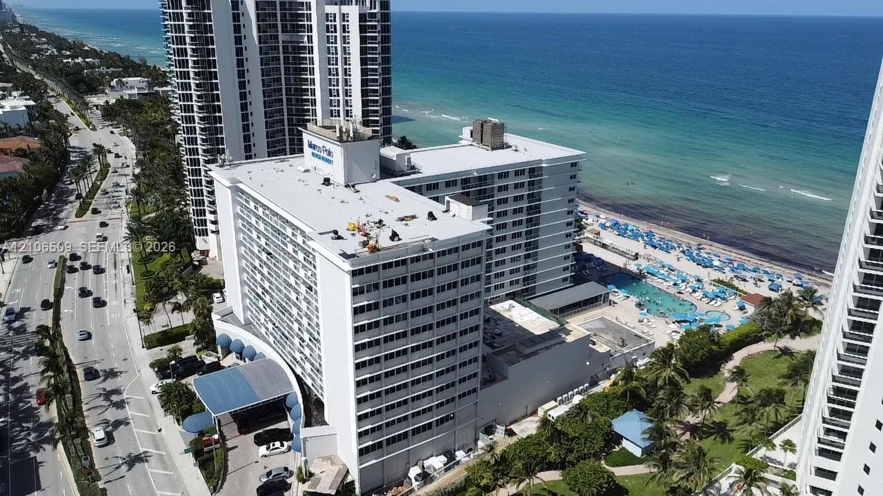 High-rise coastal property featuring direct oceanfront access, an outdoor swimming pool, a porte-cochere entryway, and mature palm landscaping