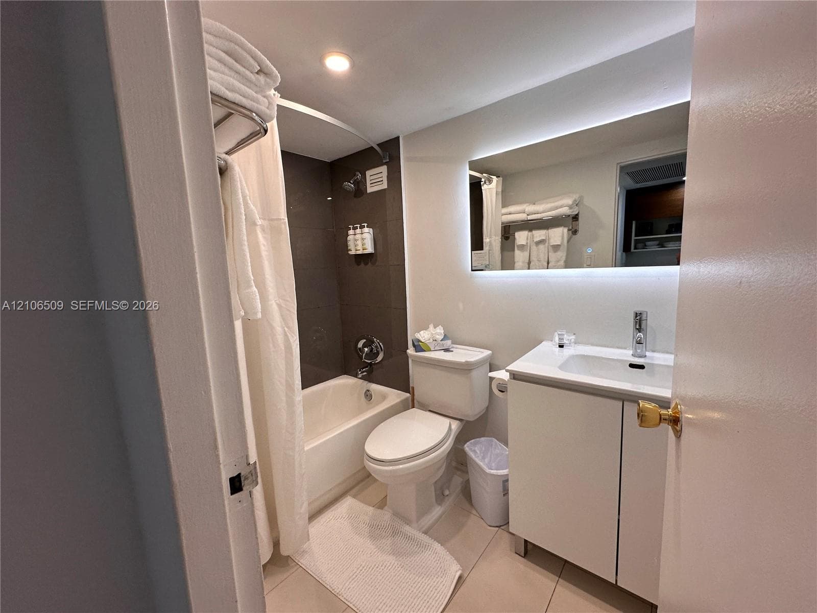 Bathroom featuring a white vanity with an integrated sink, polished chrome faucet, back-lit rectangular mirror, and a shower-tub combination with dark wall tile
