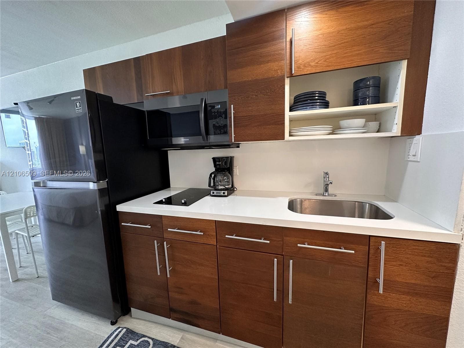 Kitchen featuring wood-finish flat-panel cabinetry, white solid-surface countertops, integrated stainless steel sink, black microwave, and black refrigerator