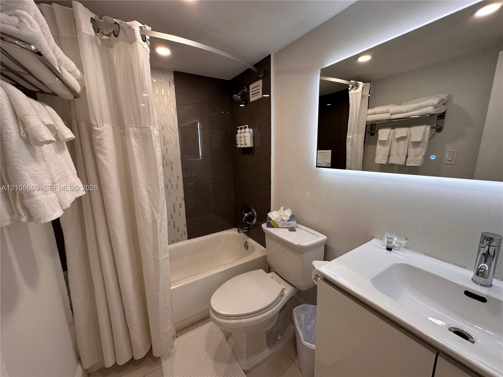 Bathroom featuring an integrated vanity with a rectangular sink, wall-mounted illuminated mirror, and a shower-tub combination with tiled surround and metal hardware