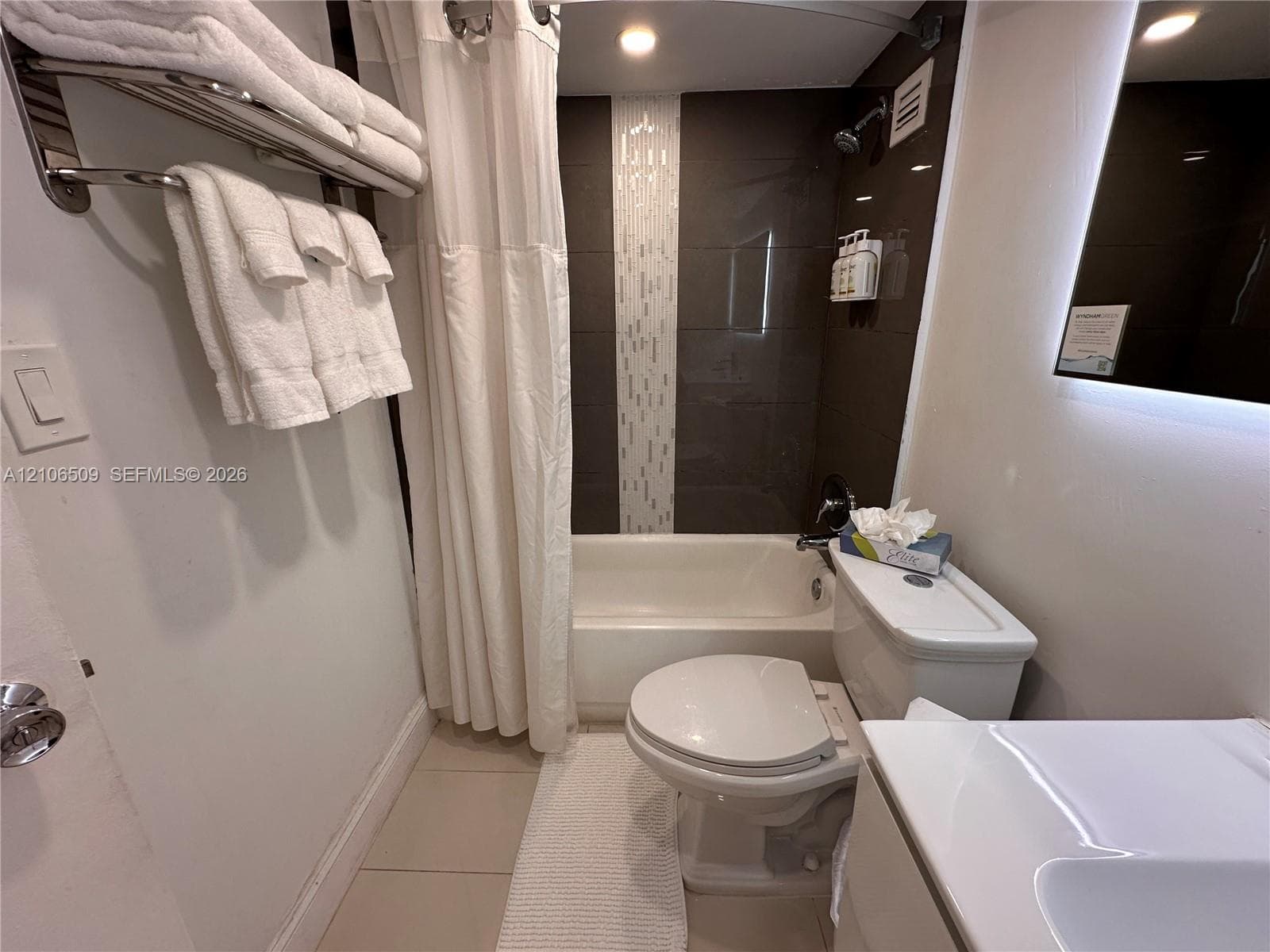 Bathroom featuring a recessed tub with a vertical decorative tile inlay, wall-mounted towel rack, LED-backlit mirror, and neutral-toned floor tile