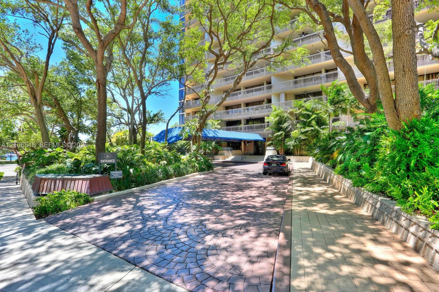 Circular paved driveway featuring stamped pattern detailing, covered porte-cochère, multi-story building exterior, mature tree canopy, and landscaped greenery
