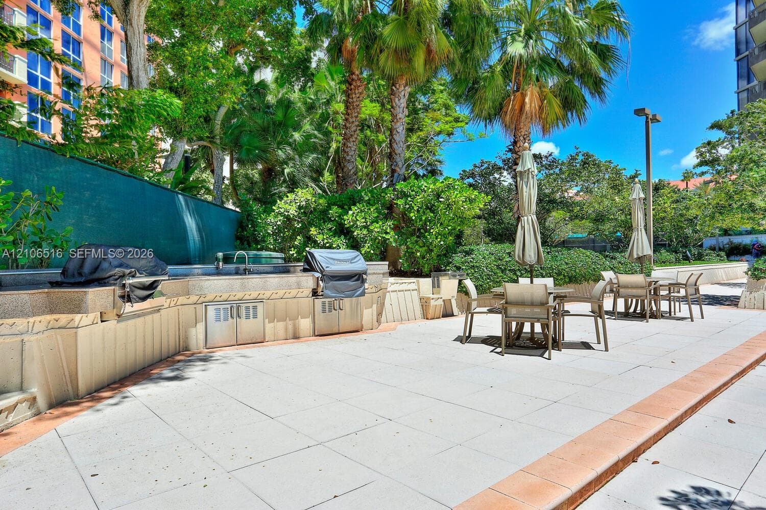 Outdoor common kitchen area featuring a stone-topped counter, built-in stainless steel cabinetry, and a sink, set within a tiled patio space surrounded by mature palm trees and greenery