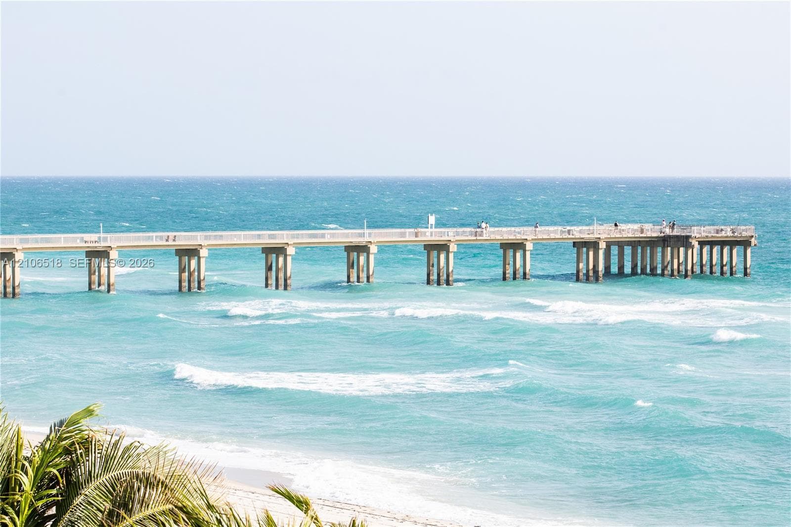 Expansive pier with white structural railing extending over turquoise ocean waters, complemented by coastal sandy shoreline and tropical palm foliage