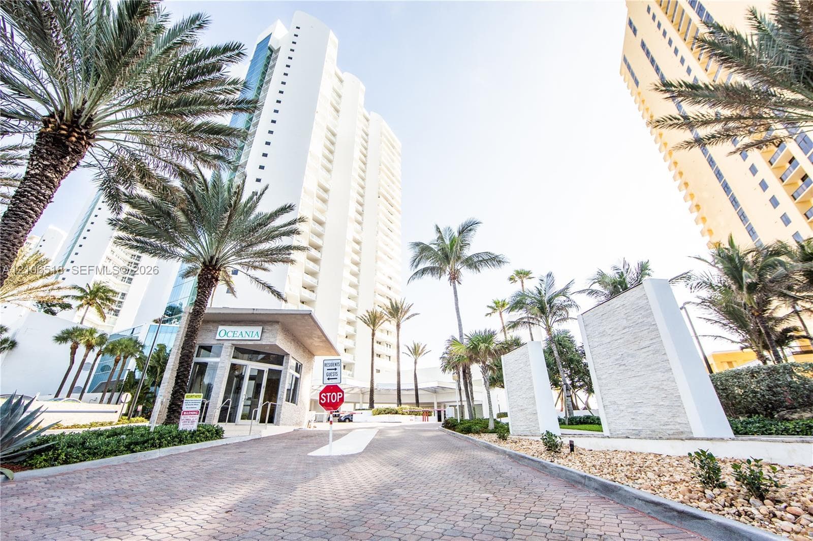 High-rise exterior featuring a paved arrival courtyard, stacked-stone entrance pillars, and extensive mature palm tree landscaping