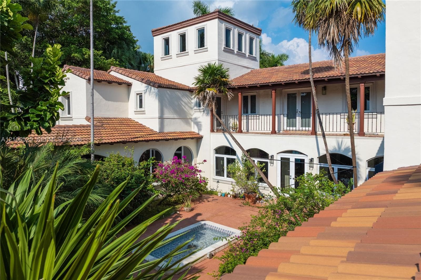 Mediterranean-style exterior featuring a stucco facade, clay tile roof, central courtyard, second-story balcony with metal railing, and a built-in stone-accented fountain