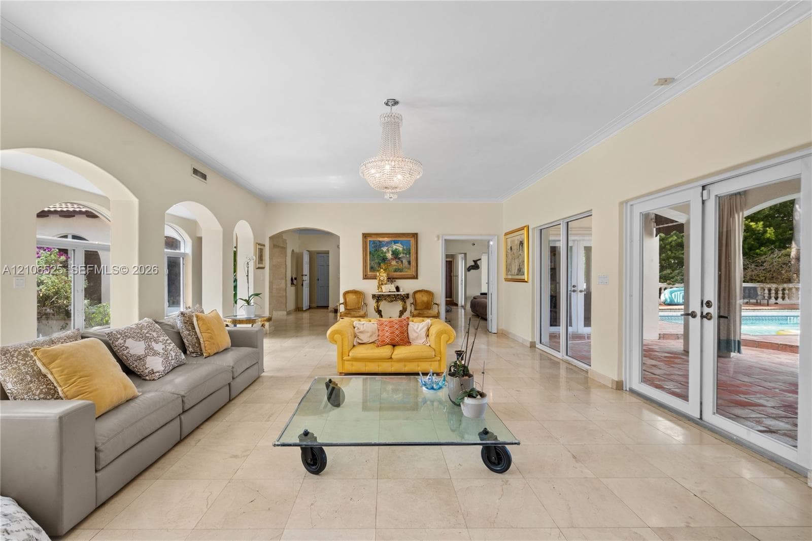 Spacious living area featuring polished stone flooring, arched architectural details, a central crystal chandelier, and two sets of glass-paned double doors