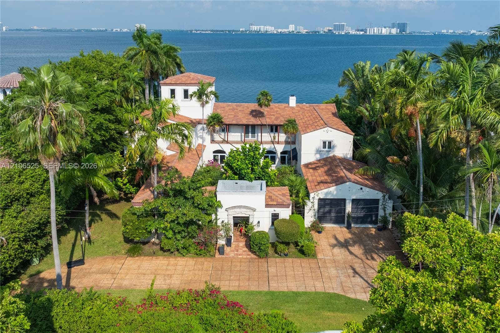 Mediterranean-style estate featuring a terracotta tile roof, multi-story tower architecture, a paved motor court, and an attached two-car garage, situated on a waterfront lot with mature palm landscaping