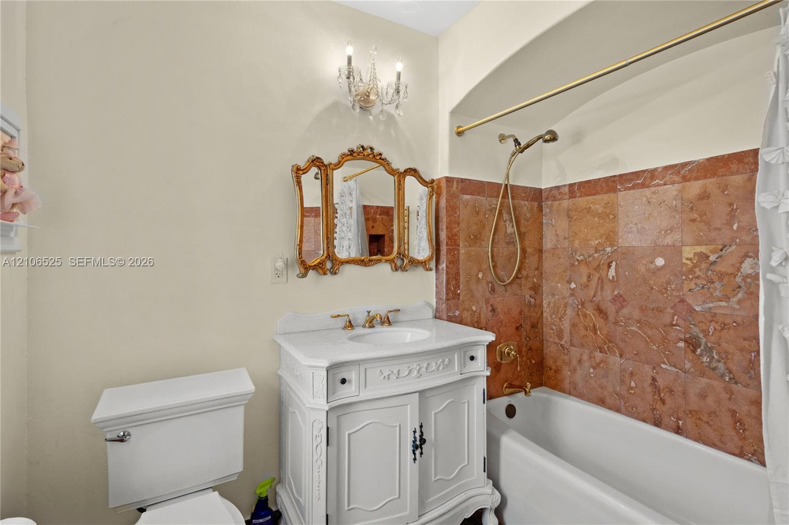 Bathroom featuring a white vanity with carved detailing, stone tile shower surround, brass-finish fixtures, gold-toned ornamental mirror, and a crystal-accented wall-mounted light fixture