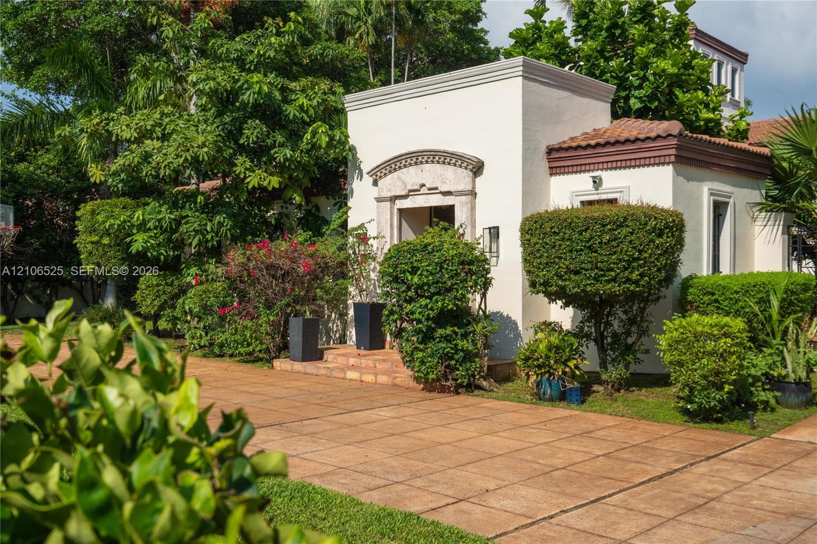 Stucco exterior featuring arched stone entryway detail, tile roof, and expansive terracotta-style paved patio area surrounded by mature landscaping