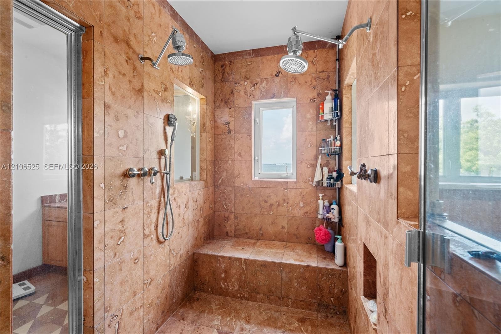 Walk-in shower enclosure featuring floor-to-ceiling stone tile, a built-in tiled bench, dual overhead rainfall showerheads, a handheld shower fixture, and recessed wall shelving