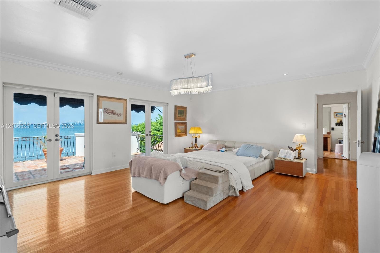 Spacious interior featuring wood-finish flooring, dual sets of French doors, crown molding, a decorative crystal chandelier, and a balcony with water scenery