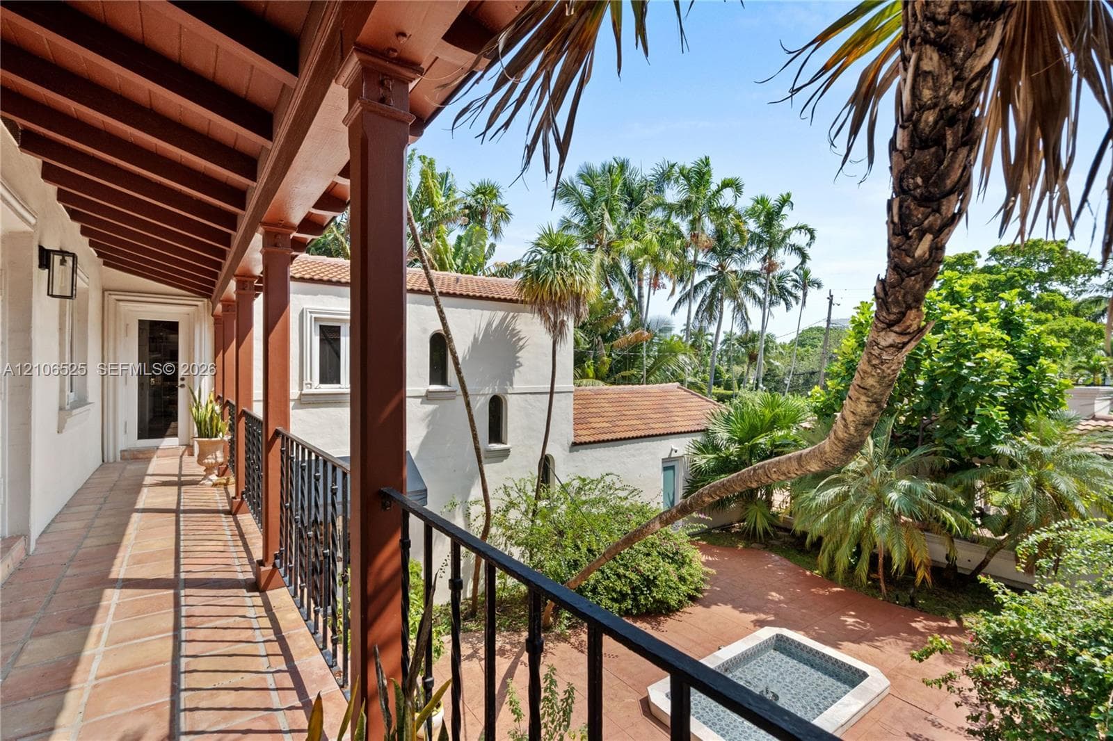 Covered balcony featuring exposed wood beam ceilings, wrought-iron railings, and tile flooring, overlooking a paved courtyard with a built-in stone fountain