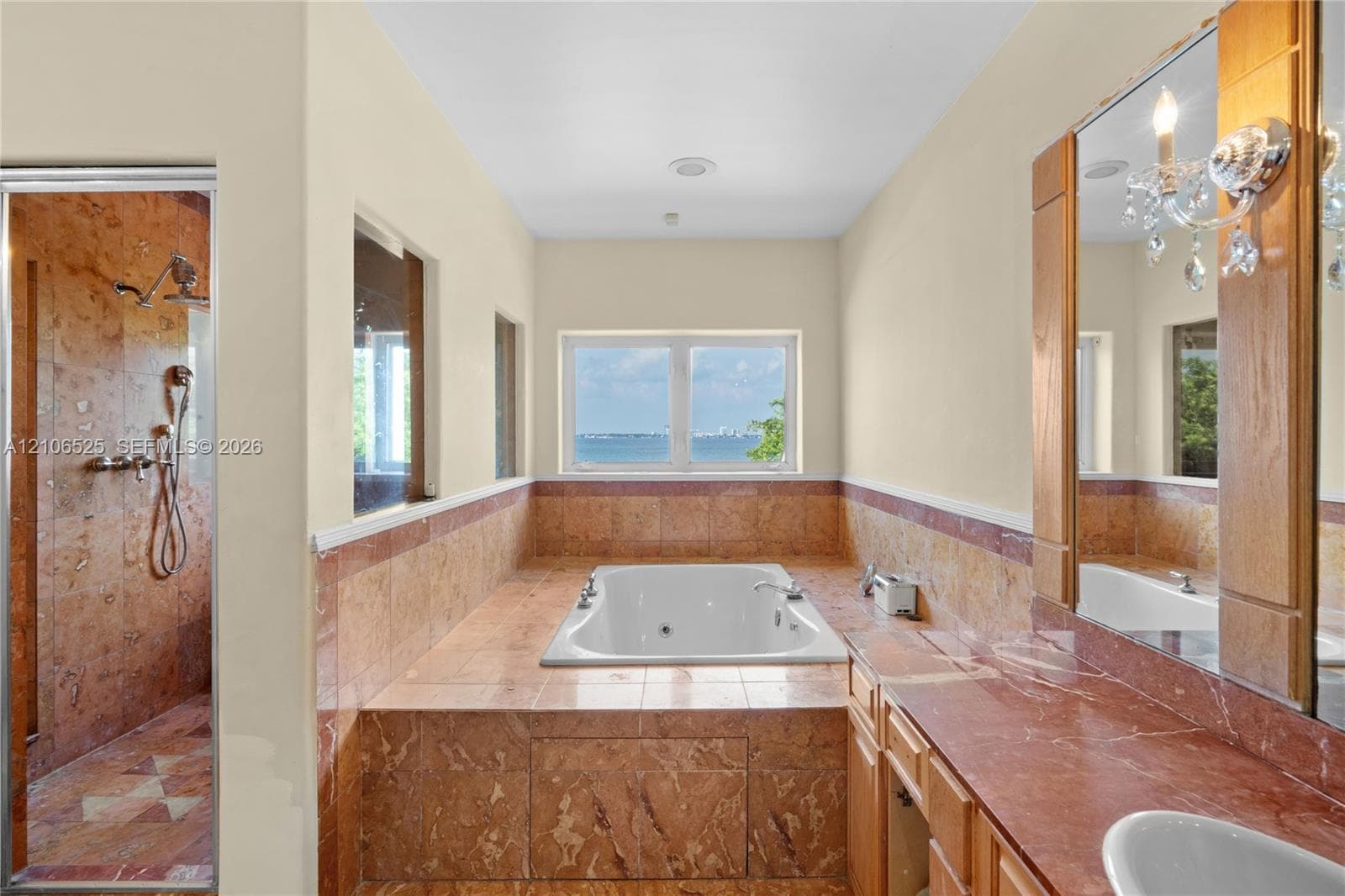 Bathroom featuring a built-in soaking tub, natural stone tile surround, walk-in shower with stone tile walls, stone-topped vanity, and wall-mounted light fixtures