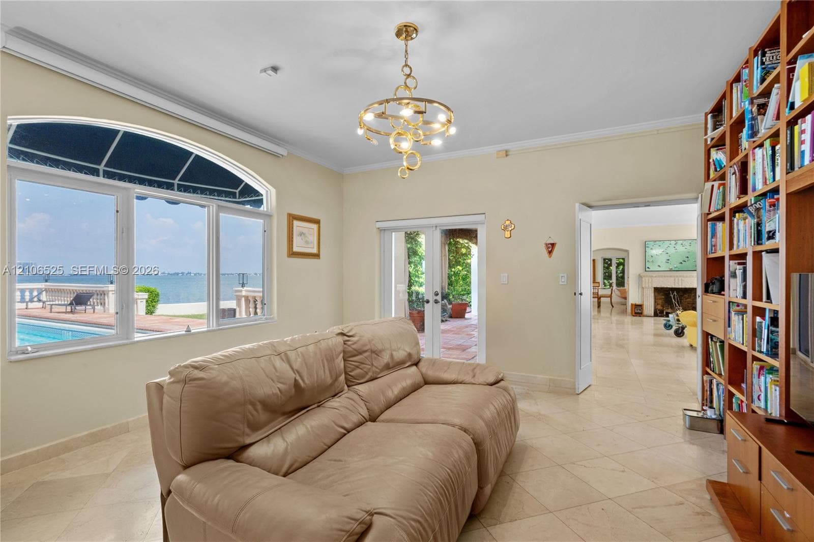 Large arched window with water and pool expanse, built-in floor-to-ceiling wood shelving, gold-tone contemporary chandelier, and light-colored stone flooring