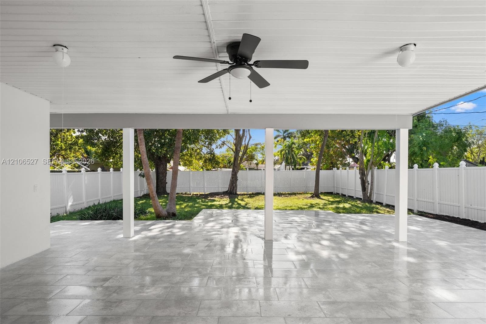 Covered patio featuring stone-finish flooring, a ceiling fan, dual overhead light fixtures, and a white vinyl privacy fence surrounding the rear grounds