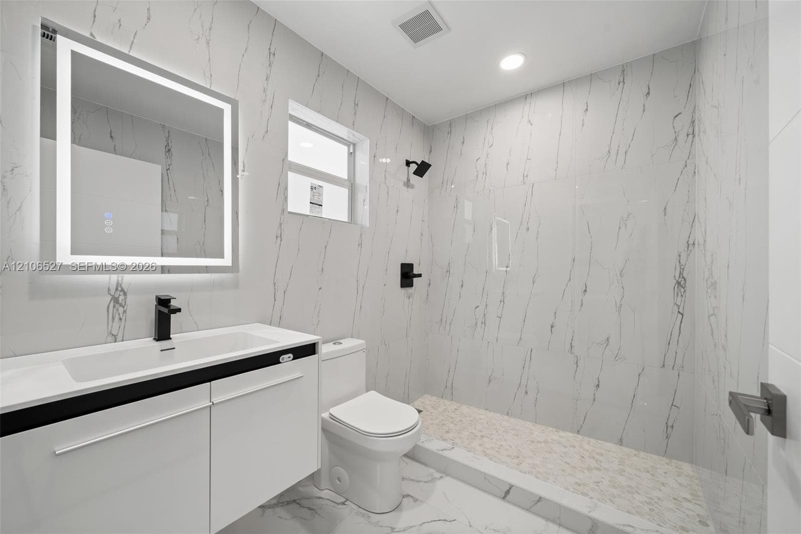 Bathroom featuring full-height stone-finish wall tile, walk-in shower with pebble-stone flooring, integrated LED vanity mirror, white cabinetry with a built-in sink, and matte black fixtures