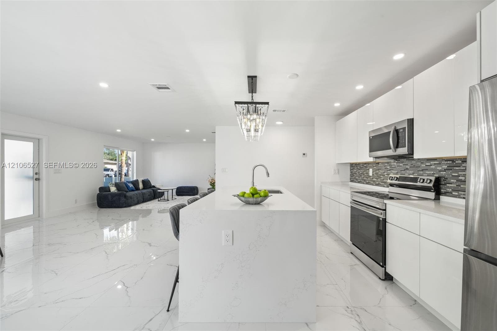 Kitchen featuring white flat-panel cabinetry, a central island with a solid-surface waterfall countertop, stainless steel appliances, a glass-tile backsplash, and polished stone-finish flooring
