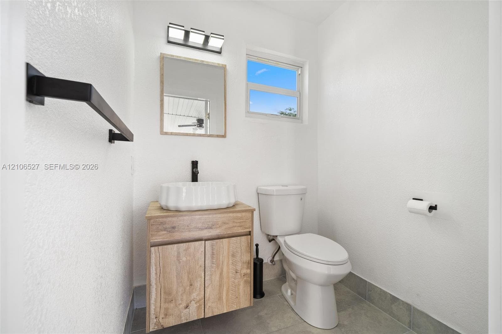 Wood-finish vanity cabinet, rectangular vessel sink, matte black wall-mounted faucet, framed wall mirror, and neutral tile flooring