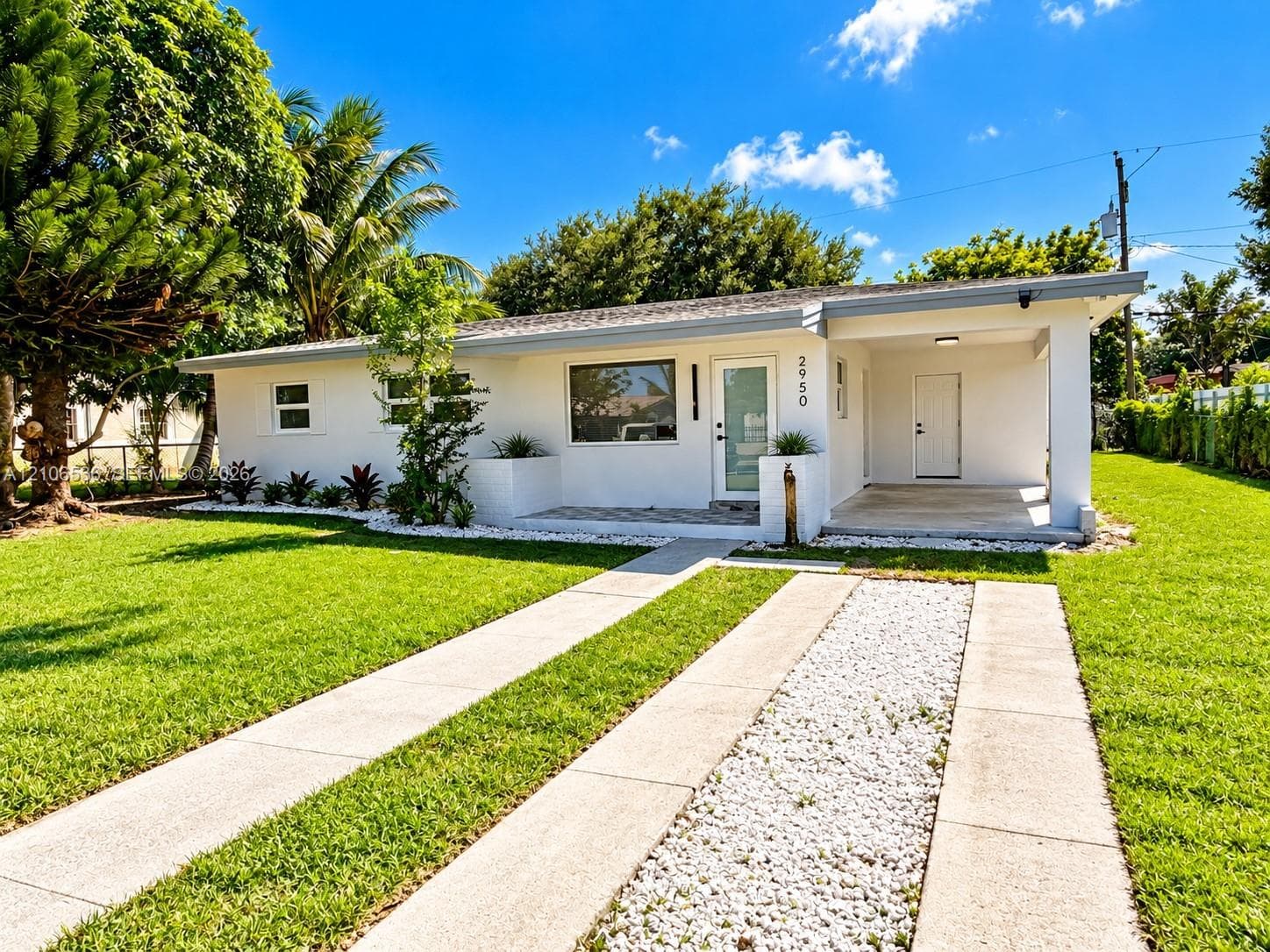 Single-story exterior featuring white stucco siding, an attached carport, a concrete ribbon driveway with a central stone aggregate strip, and manicured landscaping