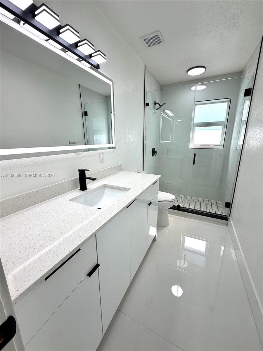 Modern vanity featuring white cabinetry, quartz countertop with an integrated basin, and a matte black faucet, illuminated by an LED-backlit mirror and five-light fixture, paired with a glass-enclosed shower featuring matte black hardware and light-toned flooring