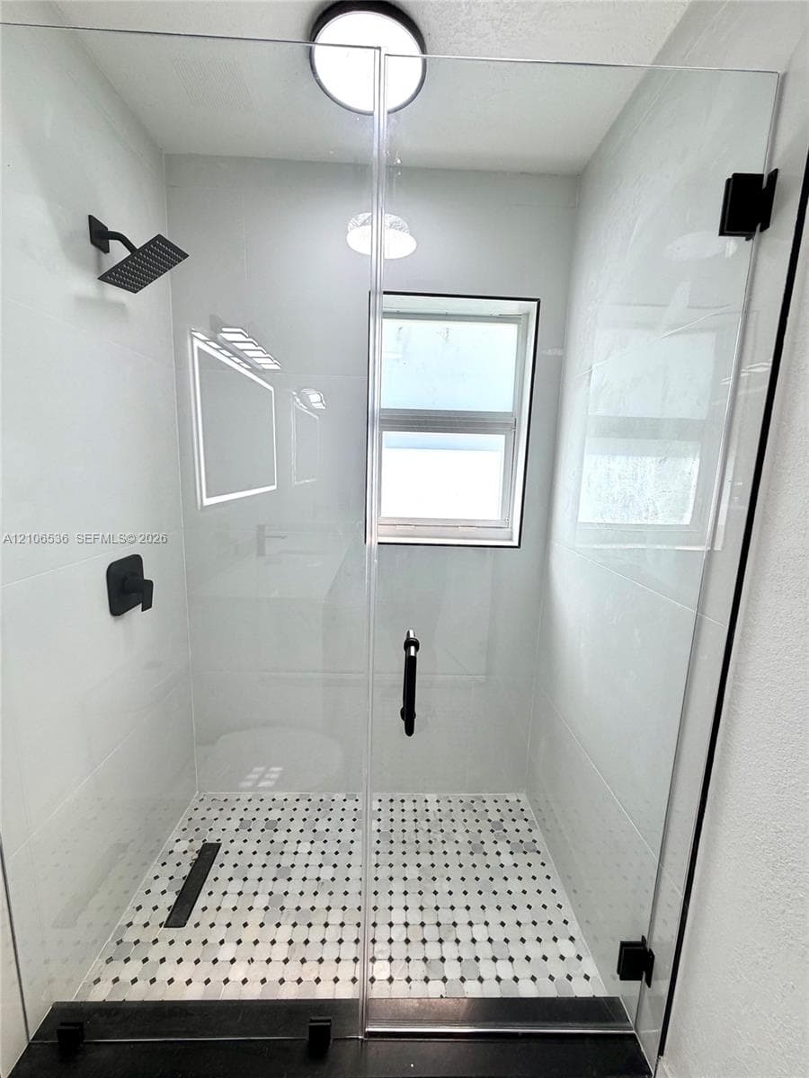 Walk-in shower featuring a frameless glass door, matte black fixtures, square rain-style showerhead, large-format wall tiling, and decorative mosaic floor tile with a linear drain