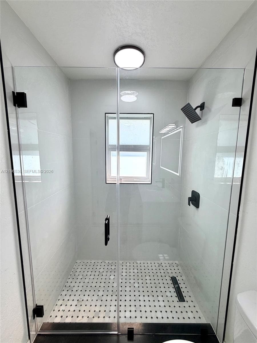 Walk-in shower featuring a frameless glass enclosure, matte black fixtures, white tiled walls, patterned floor tiling, and a recessed ceiling light