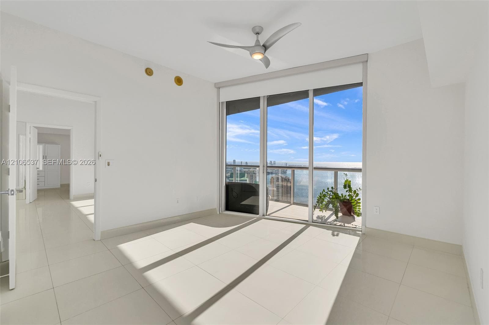 Floor-to-ceiling glass sliding doors opening to a private balcony, polished tile flooring, three-blade ceiling fan with integrated lighting, and panoramic water and city skyline outlooks