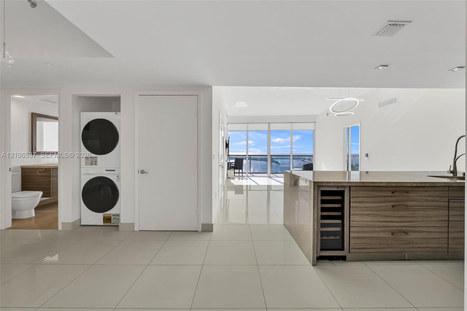 Open-concept interior featuring large-format tile flooring, stacked laundry unit, built-in wine cooler, wood-finish cabinetry, and floor-to-ceiling windows