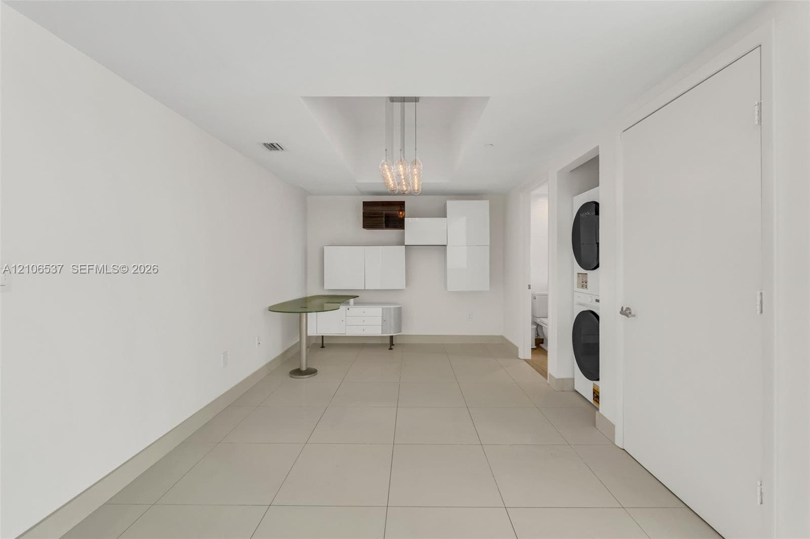 Modern utility space featuring large-format tile flooring, built-in glossy white cabinetry, an integrated stone-surface workstation, a tiered pendant light fixture, and a stacked washer and dryer unit