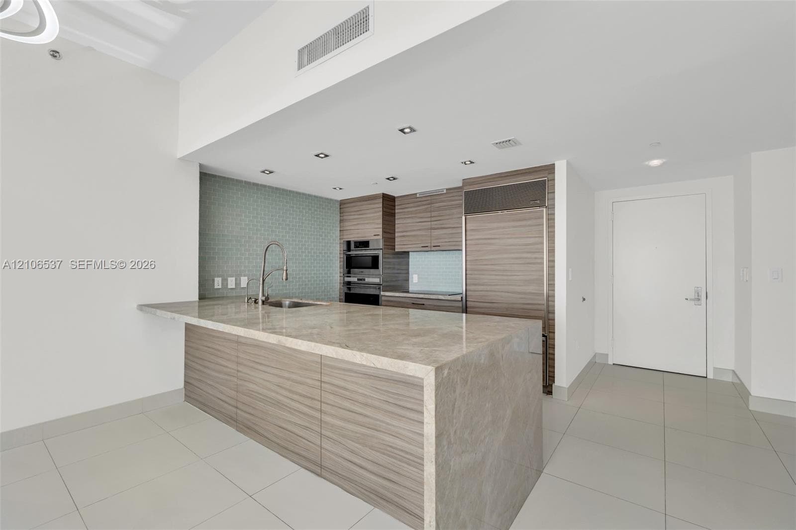 Kitchen featuring a waterfall-edge stone countertop, wood-finish cabinetry, built-in stainless steel refrigerator, recessed lighting, and patterned tile backsplash
