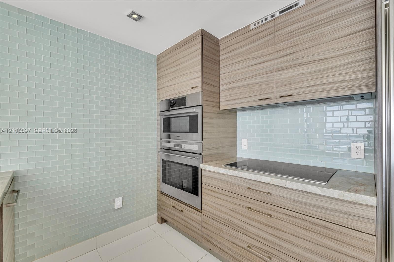Modern kitchen featuring wood-finish cabinetry, light blue subway tile backsplash, built-in stainless steel double oven, and integrated cooktop