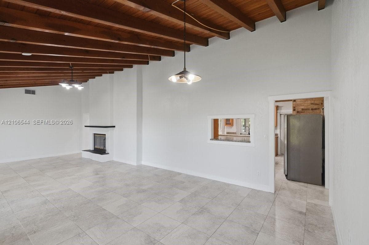 Vaulted ceiling with exposed wood beams, neutral tile flooring, fireplace with a black mantel, and two hanging pendant light fixtures