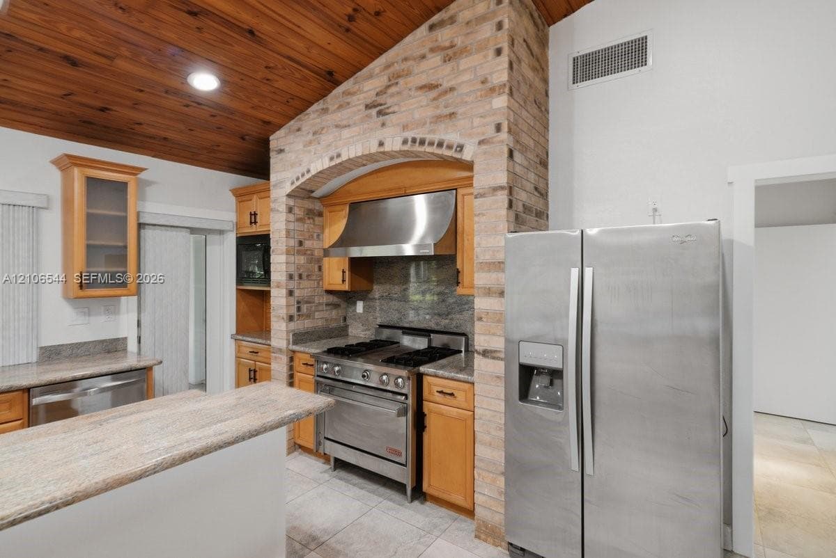 Kitchen featuring vaulted wood-plank ceiling, natural stone countertops, custom brick archway, stainless steel range with matching hood, and warm-toned cabinetry