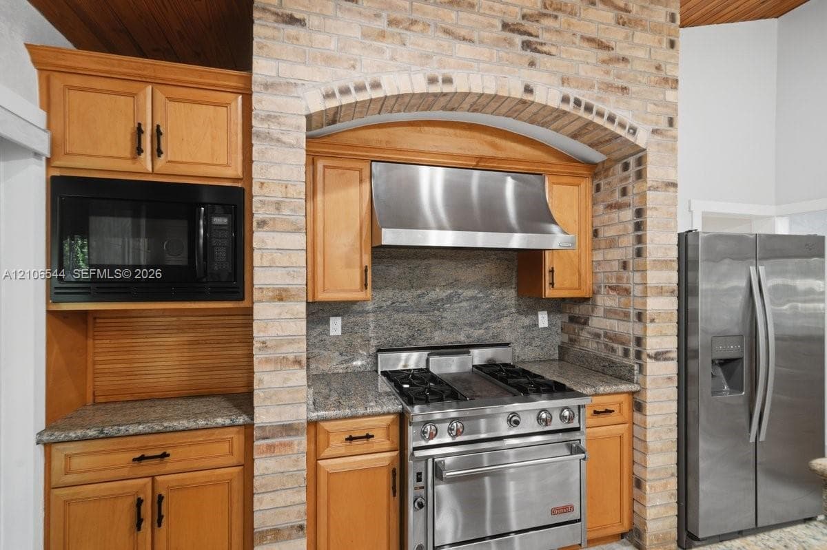 Kitchen featuring custom wood cabinetry, a stone-surround recessed cooktop area with an arched brick header, stainless steel professional-grade range, stone countertops, and a tongue-and-groove wood ceiling